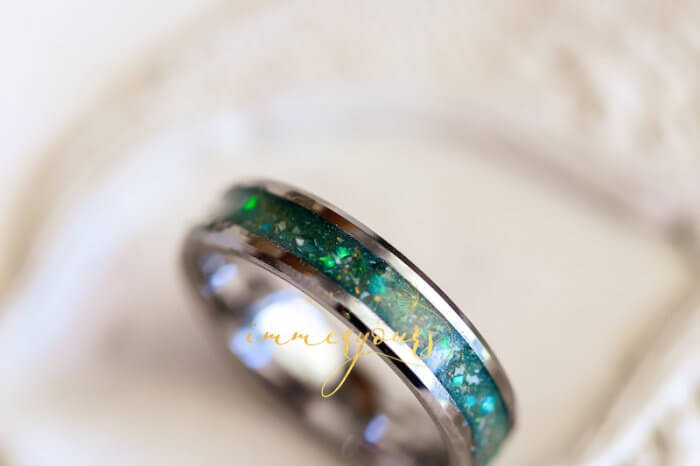 A close-up of a tungsten memorial ash ring with a green inlay. The inlay features a blend of vibrant green and iridescent flakes, creating a shimmering effect.