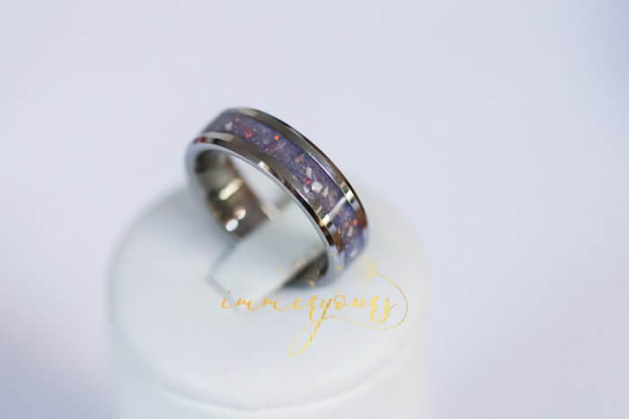 A tungsten memorial ash ring displayed on a white stand. The ring has a purple inlay with opal flakes, creating a striking and elegant design.